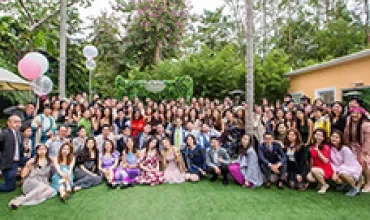 Large group of people posing outdoors at a gathering with greenery and balloons.